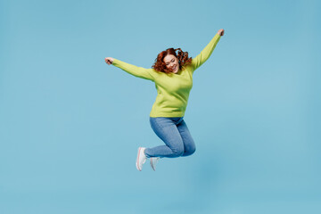 Young overjoyed excited chubby overweight plus size big fat fit woman wear green sweater jump high with outstretched hands isolated on plain blue background studio portrait. People lifestyle concept.