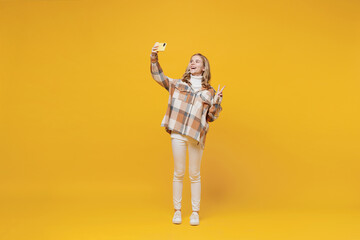 Full body little blonde fun smiling kid girl 13-14 years wearing checkered shirt doing selfie shot on mobile cell phone post photo on social network show v-sign isolated on plain yellow background