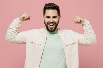 Young excited happy man 20s in trendy jacket shirt showing biceps muscles on hand demonstrating strength power isolated on plain pastel light pink background studio portrait. People lifestyle concept.