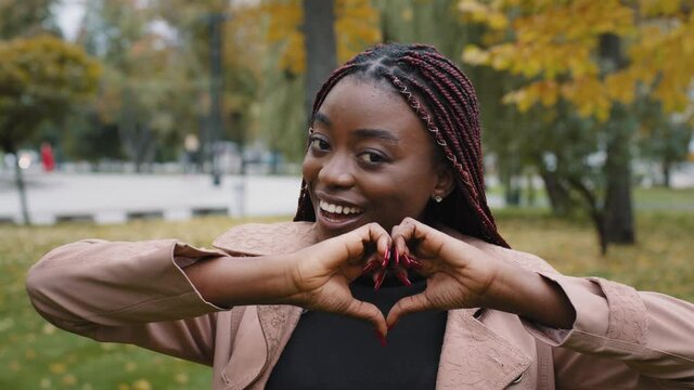 Smiling young African American woman volunteer showing hands sign heart shape looking at camera in park on autumn day. Healthy heart health life insurance, love and charity, voluntary social work