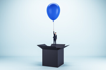 Abstract image of young european businessman flying on blue balloon out of black paper box on light background. Success, business and happiness concept.
