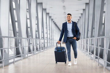Portrait of young middle eastern man walking in airport terminal with luggage - obrazy, fototapety, plakaty