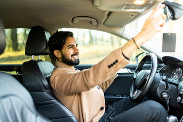 Confident young man wearing glasses adjusting rear view mirror driving alone in luxury car, back view, reflection