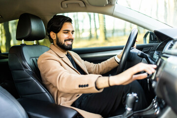 Handsome indian young man in denim shirt pressing touchscreen on car multimedia panel, switching shifting radio station.