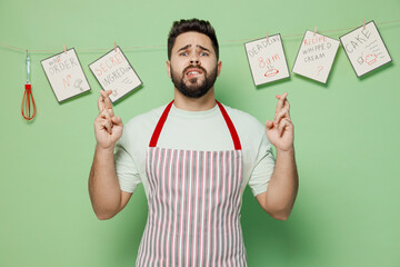 Young male chef confectioner baker man in striped apron waiting for special moment, keeping fingers crossed, making wish isolated on plain pastel light green background studio. Cooking food concept.