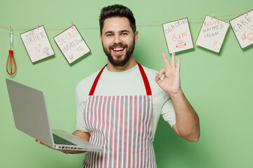 Young male chef confectioner baker man in striped apron hold use work on laptop pc computer show ok okay gesture isolated on plain pastel light green background studio portrait. Cooking food concept.