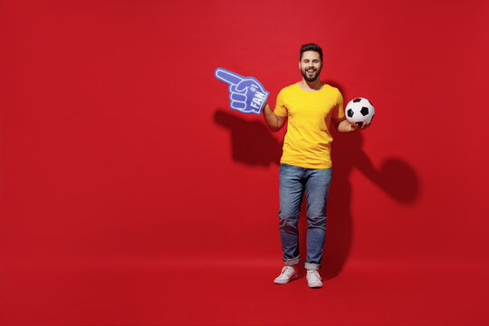 Full size body length young man football fan in yellow t-shirt hold soccer ball point fan foam glove finger on workspace area copy space mock up isolated on plain dark red background studio portrait.