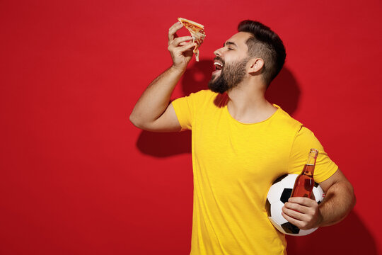 Satisfied hungry young bearded man football fan in yellow t-shirt cheer up support favorite team hold soccer ball beer bottle eat italian pizza isolated on plain dark red background studio portrait.