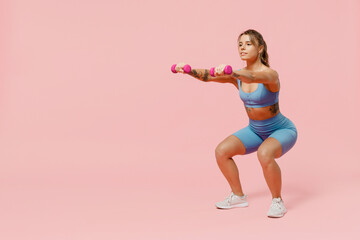 Full size young sporty athletic fitness trainer instructor woman wear blue tracksuit spend time in home gym hold dumbbells do squats isolated on plain light pink background. Workout sport concept