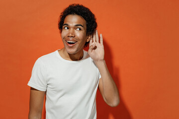 Curious nosy smilimg vivid shocked joyful amazed young black curly man 20s years old wears white t-shirt try to hear you overhear listening isolated on plain pastel orange background studio portrait.