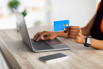 Closeup of black woman holding credit card and using laptop, making e-payments on computer, shopping online from home