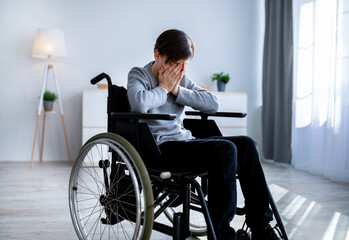 Depression in disabled teenagers. Unhappy handicapped teen boy in wheelchair crying at home