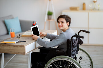 Positive disabled teenager in wheelchair using tablet computer, having online lesson or webinar at home