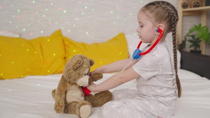 little child listens teddy bear with stethoscope, kid girl plays doctor while sitting on bed, childhood dream becoming nurse, veterinarian, baby helping animals, happy family, having fun playing toys