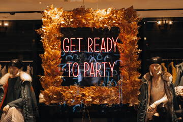 Neon shiny holiday signboard on boutique showcase with text Get ready for party and female mannequins