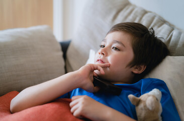 Happy boy lying on sofa watching  cartoon, School Kid sitting alone on couch holding remote control and watching TV, Smiling Child relaxing in living room after back from school.
