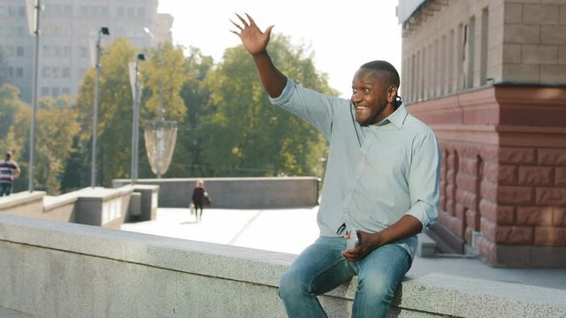 Elderly man in casual clothes waving hand affably. Retirement african american person, businessman or freelancer sitting outdoors holding smartphone looking at phone screen greeting friend