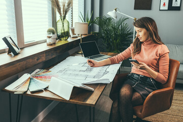 Young woman working on a project. Female designer in office working on architects project. Female architect working at home. She looking at blueprint. Multitasking of young female entrepreneur at work