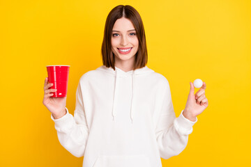 Photo of cheerful lady hold plastic cup tennis ball beer pong wear white hoodie isolated yellow...