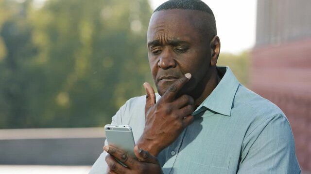 Elderly African man sitting outdoors holding smartphone experiences difficulties with wireless modern gadget usage, wrong password, need device repair or help with app understanding concept