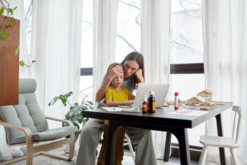 Weary mom leaning on hand while touching head of sick son browsing netbook at table with medicine bottles at home