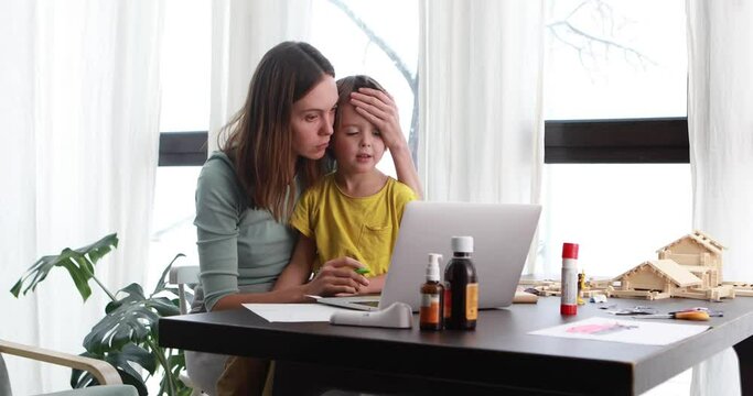 Weary mom leaning on hand while touching head of sick son browsing netbook at table with medicine bottles at home