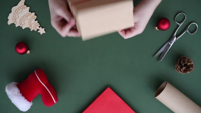 Female hands pack a gift for Christmas ans New Year winter holidays in eco-friendly materials on green background.Craft paper box tied with red ribbon bow.Santa's shoe, red balls, package, scissors.