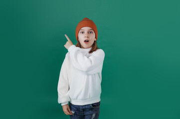 a handsome young boy in a white sweater and hat shows and looks away in surprise on an isolated green background.