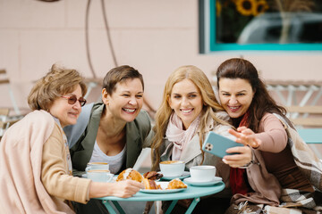 Three Caucasian pretty senior women friends sitting with coffee at table in cafe terrace and smiling to smartphone camera while taking selfie photos. Old ladies posing to phone. Pensioners resting.