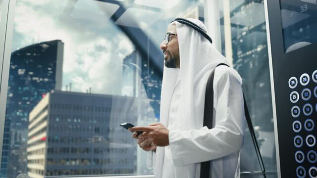 Successful Muslim Businessman in Traditional White Kandura Riding Glass Elevator to Office in Modern Business Center. Man Using Smartphone. Saudi, Emirati, Arab Businessman Concept.