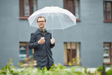 Horizontal medium shot of handsome Caucasian man wearing eyeglasses and black raincoat walking under umbrella with cup of coffee looking away