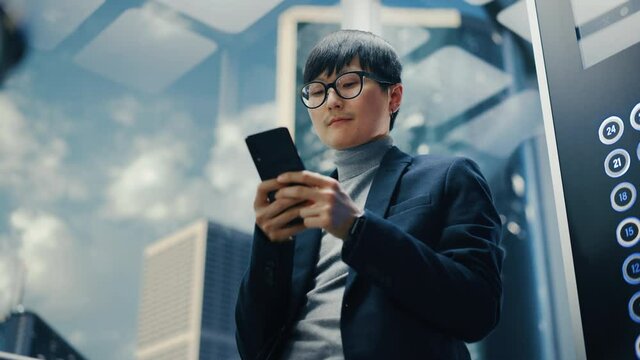 Successful Stylish Japanese Businessman Riding Glass Elevator to Office in Modern Business Center. Handsome Man Smile while Using Smartphone, Write Text Message, Check Social Media in a Lift.