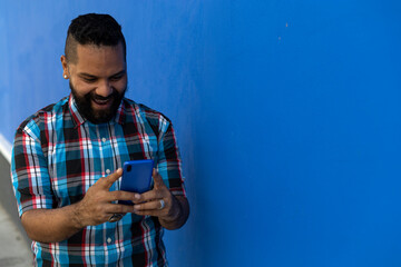 Adult male (39) with beard and plaid shirt, smiles while reading text message on his cell phone blue color, blue wall background. Copy space.