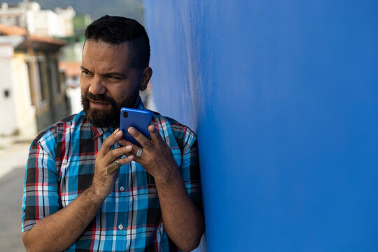 Adult man (39) with beard and checkered shirt, listens to a voice message on his cell phone blue color, city background and blue wall. Copy space.