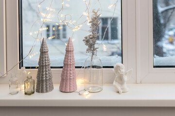 Gray and pink ceramic Christmas trees, a glass bottle with an artificial silver branch and garland decorate the windowsill for Christmas.