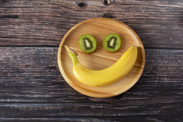 Fototapeta na wymiar Funny face made from banana and kiwi on a wooden plate on a wooden background.