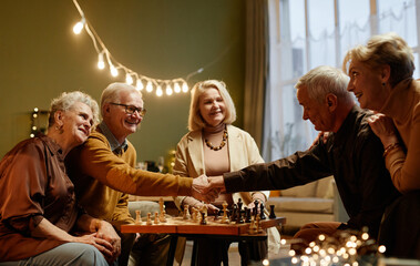 Two aged Caucasian men shaking hand after finishing chess play while three old women sitting nearby and watching