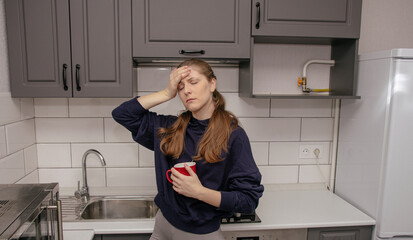 A young brunette girl stands sad and unhappy in the kitchen alone with a red mug, holds her right...