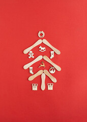 Naklejka na ściany i meble New year tree made of wooden ice cream stick and decoration baubles, reindeer, sock, a gift, cavalry and gingerbread man on red background. Creative Christmas minimal flat lay concept.