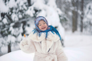 Obraz premium A girl in a white fur coat and a Russian folk blue shawl with patterns holds a lollipop cockerel on a stick in a winter snowy forest