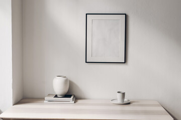 Fototapeta na wymiar Elegant minimal home office still life composition. White vase, cup of coffee, magazines. Wooden table background in sunlight. Empty black picture frame, white living room wall. Scandinavian interior.