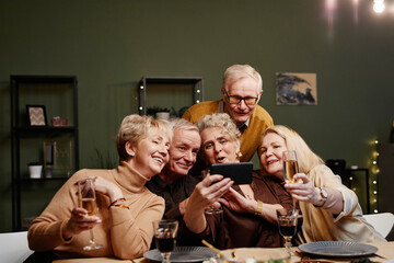 Group of five cheerful senior friends gathered at dining table for celebration, holding glasses and making selfie on smartphone camera