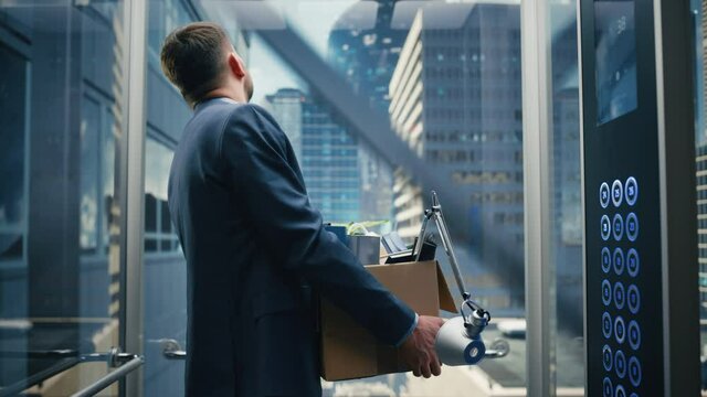 Fired Manager Going Down from Office in a Glass Elevator in Modern Business Center. Sad Specialist Laid Off, Feeling Depressed and Devastated, Holding Office Accessories and a Plant in a Cardboard Box