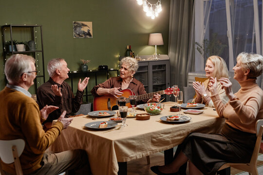 Beautiful old woman playing acoustic guitar while sitting at dining table in decorated living room and having festive dinner with her four friends