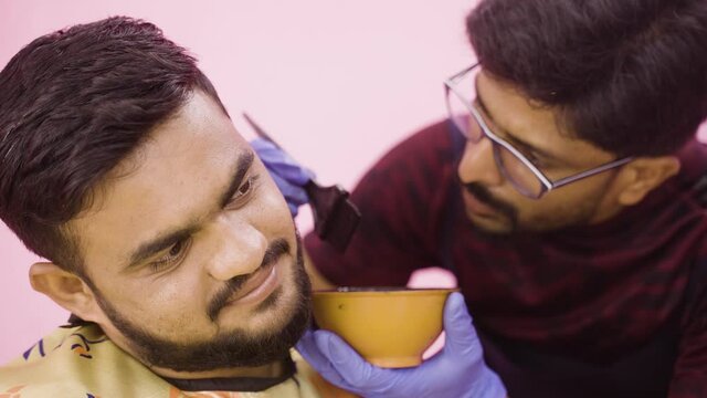 Rack focus shot, close up shot of barber applying hair dye or black henna to young man to cover white hair beard at salon - concept of hair coloring service and small business.