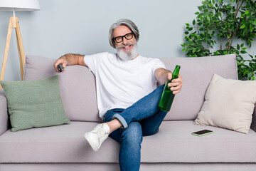 Photo of pretty relaxing retired man wear white t-shirt glasses sitting coach watching tv drinking beer indoors isolated grey color background