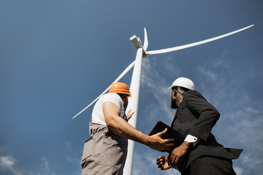 Group of multiracial coworkers examining wind turbines using modern tablet outdoors. Two men in uniform and suit talking about clean alternative energy.