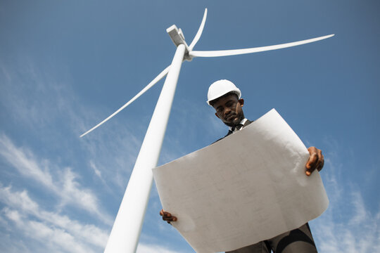 Low angle view of focused african american man standing on field with wind turbines and examining blueprints that holding in hands. Renewable and sustainable energy concept.