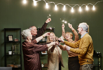 Two aged men and three women standing together in decorated with lights living room, holding sparklers in hands and clinking glasses during festive meeting