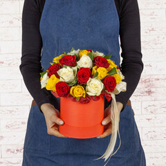 Young woman florist holding big beautiful blossoming red, white and yellow roses in red cardboard...